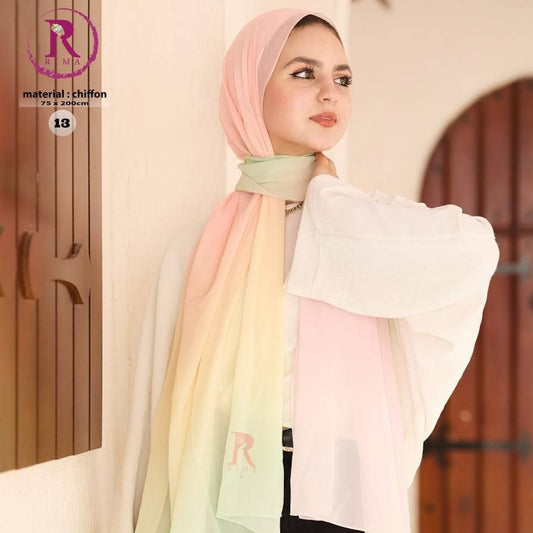A woman wearing a pastel gradient chiffon hijab scarf with pink, yellow, and green tones, paired with a white blouse and black pants, standing indoors against a cream-colored wall with wooden door frames.
